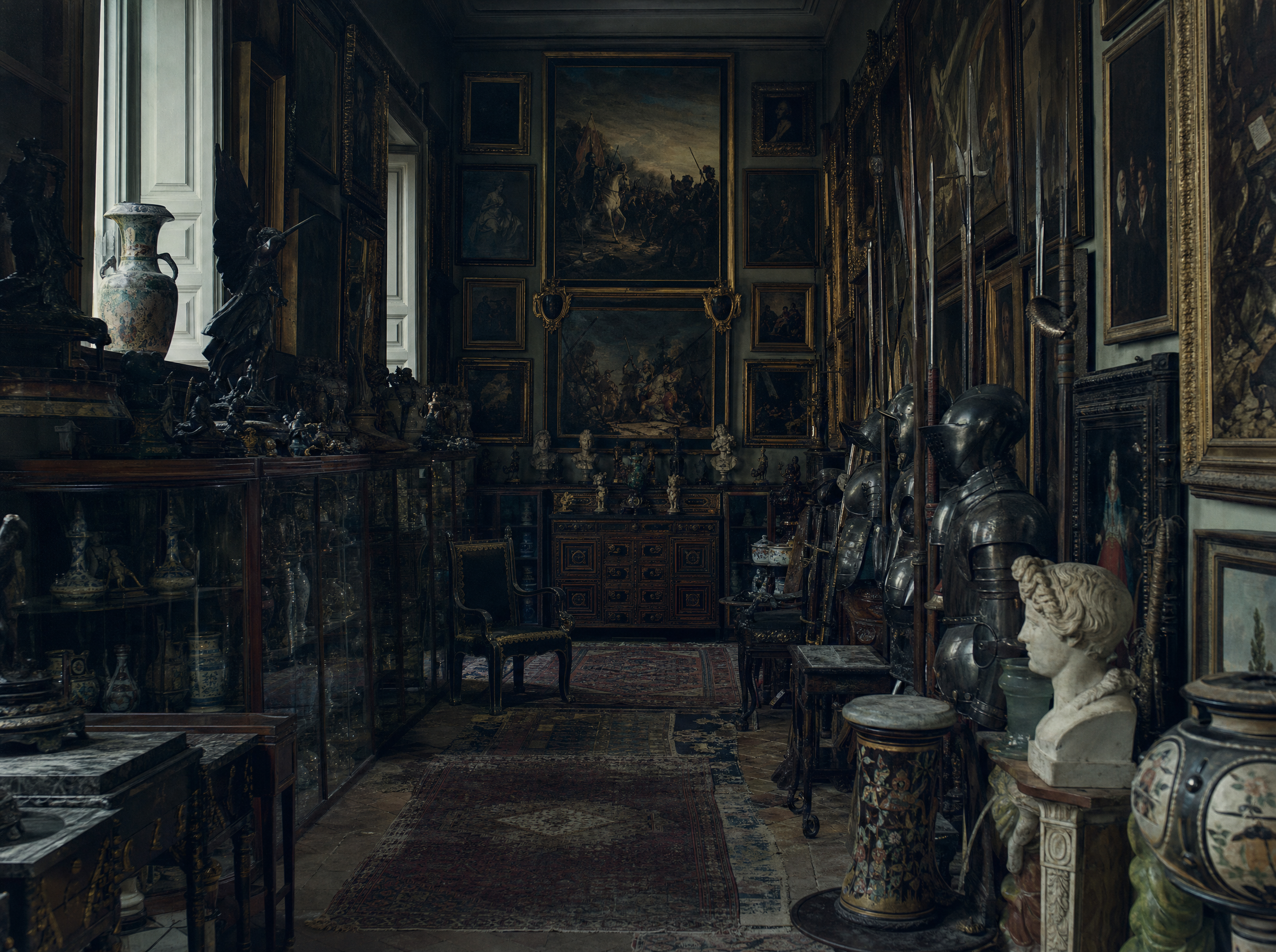 A collector's room packed with glass cabinets, ranked armour and paintings hung frame to frame