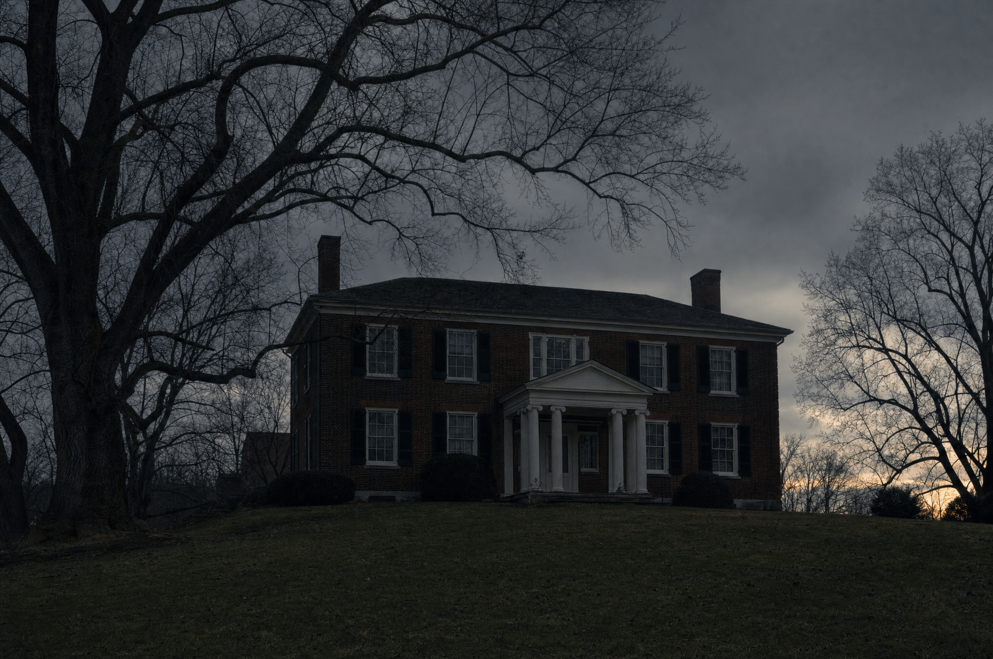 A Federal-period brick house with a portico behind a lawn at dusk, bare oaks and a low grey sky