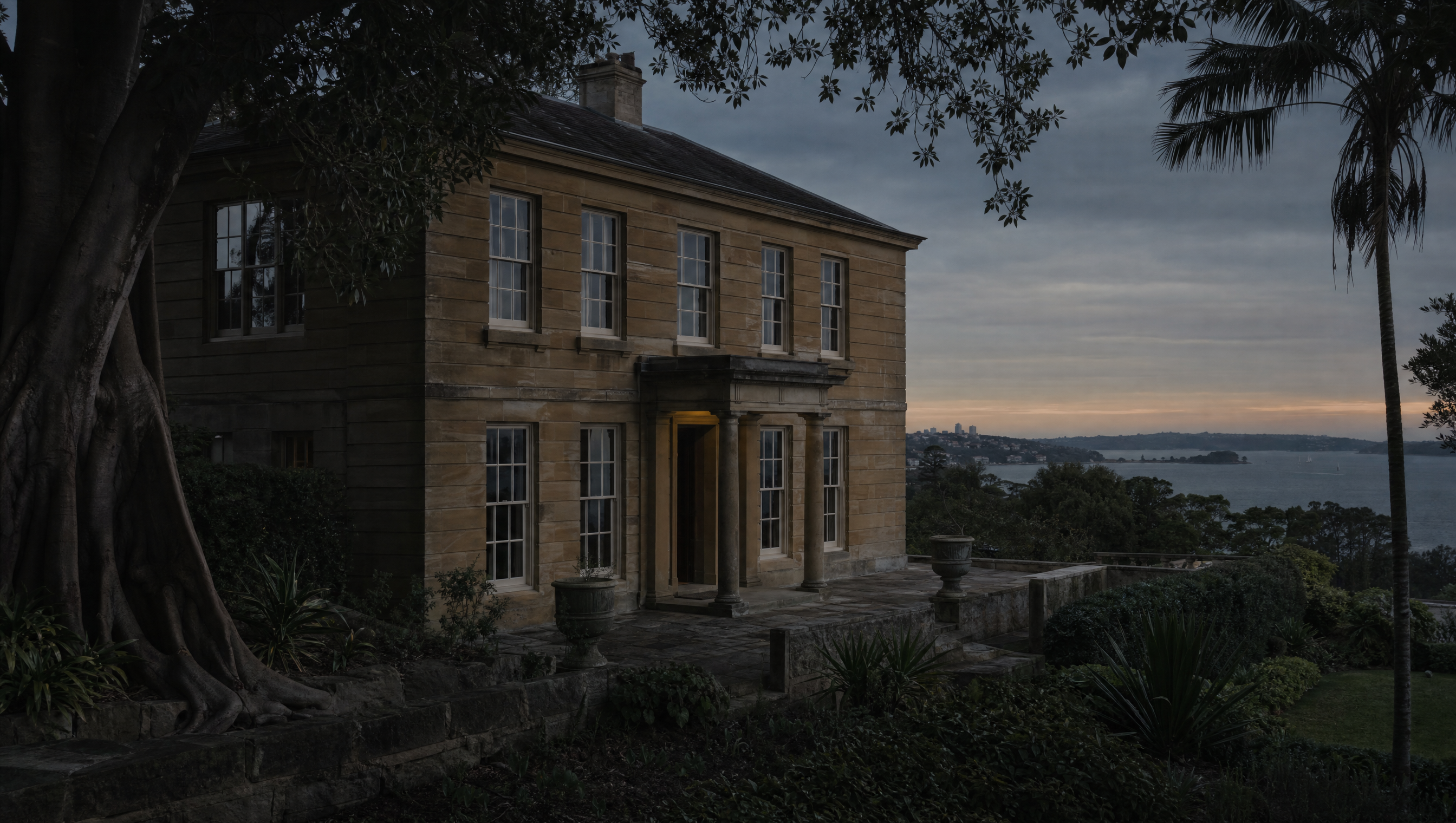 The sandstone facade of Elizabeth Bay House at dusk, seen from the terraced garden with the harbour beyond