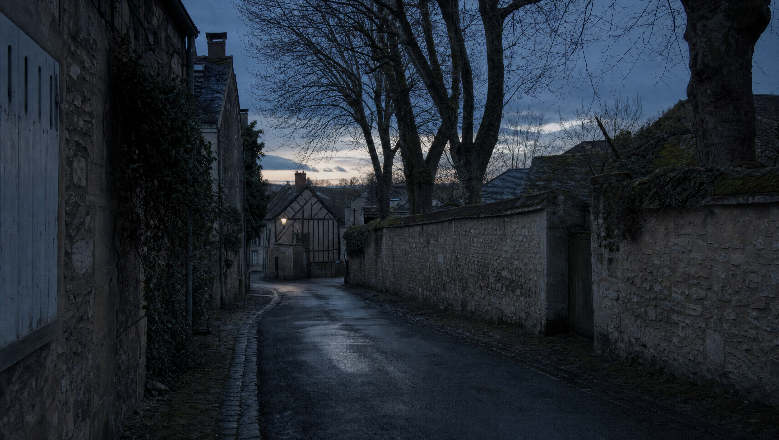 A narrow Norman village lane at dusk, rendered walls and shuttered windows under bare trees