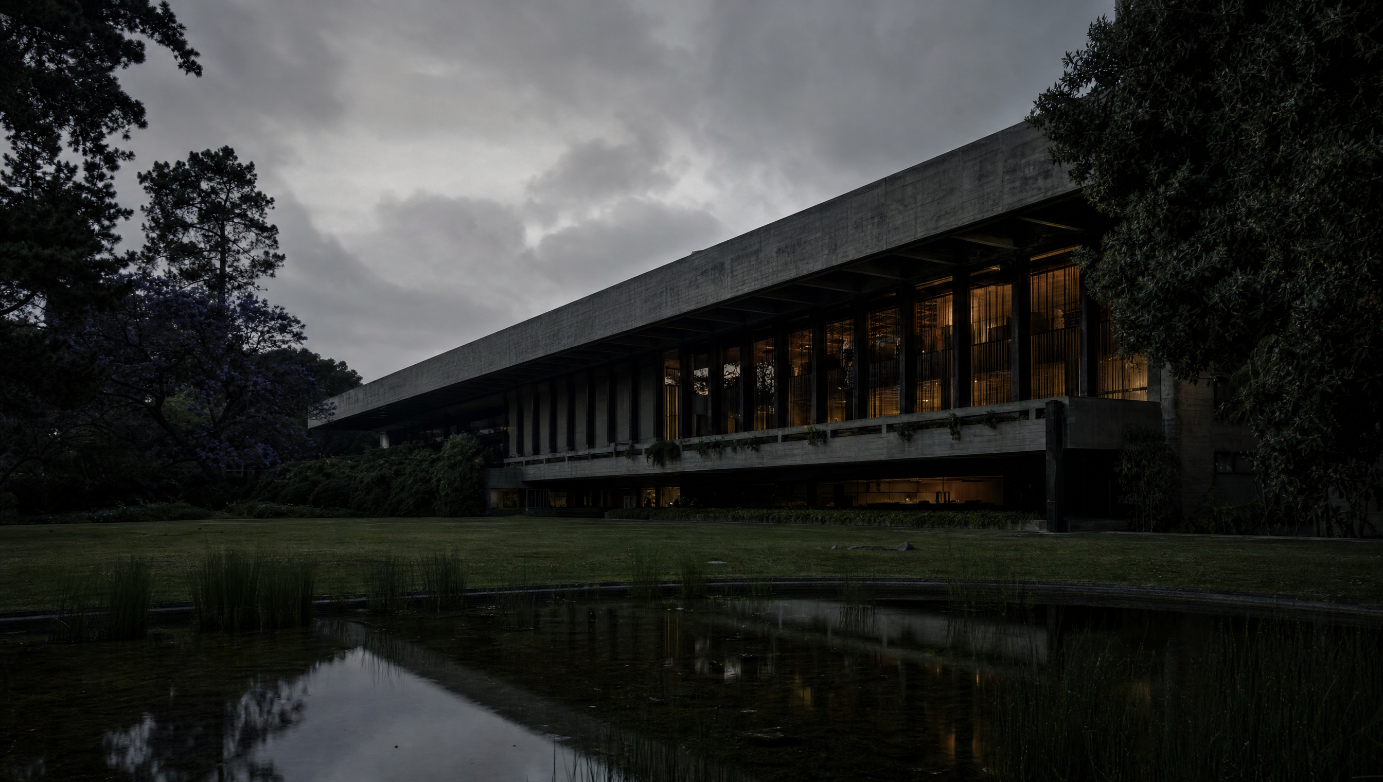 The long low concrete and granite front of the Calouste Gulbenkian Museum at dusk, its lit galleries reflected in the garden water