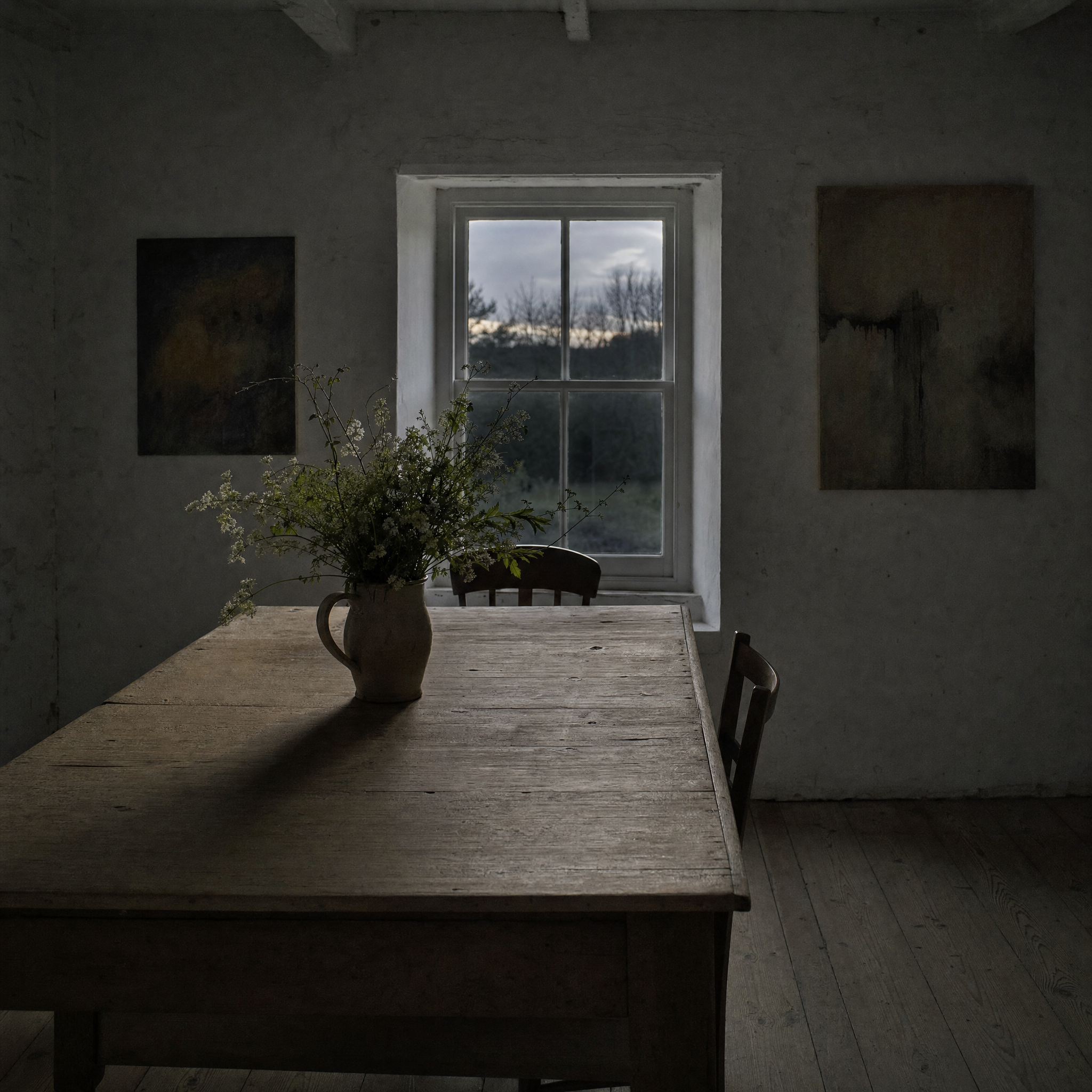 A small whitewashed farmhouse room with a plain timber table and paintings hung low