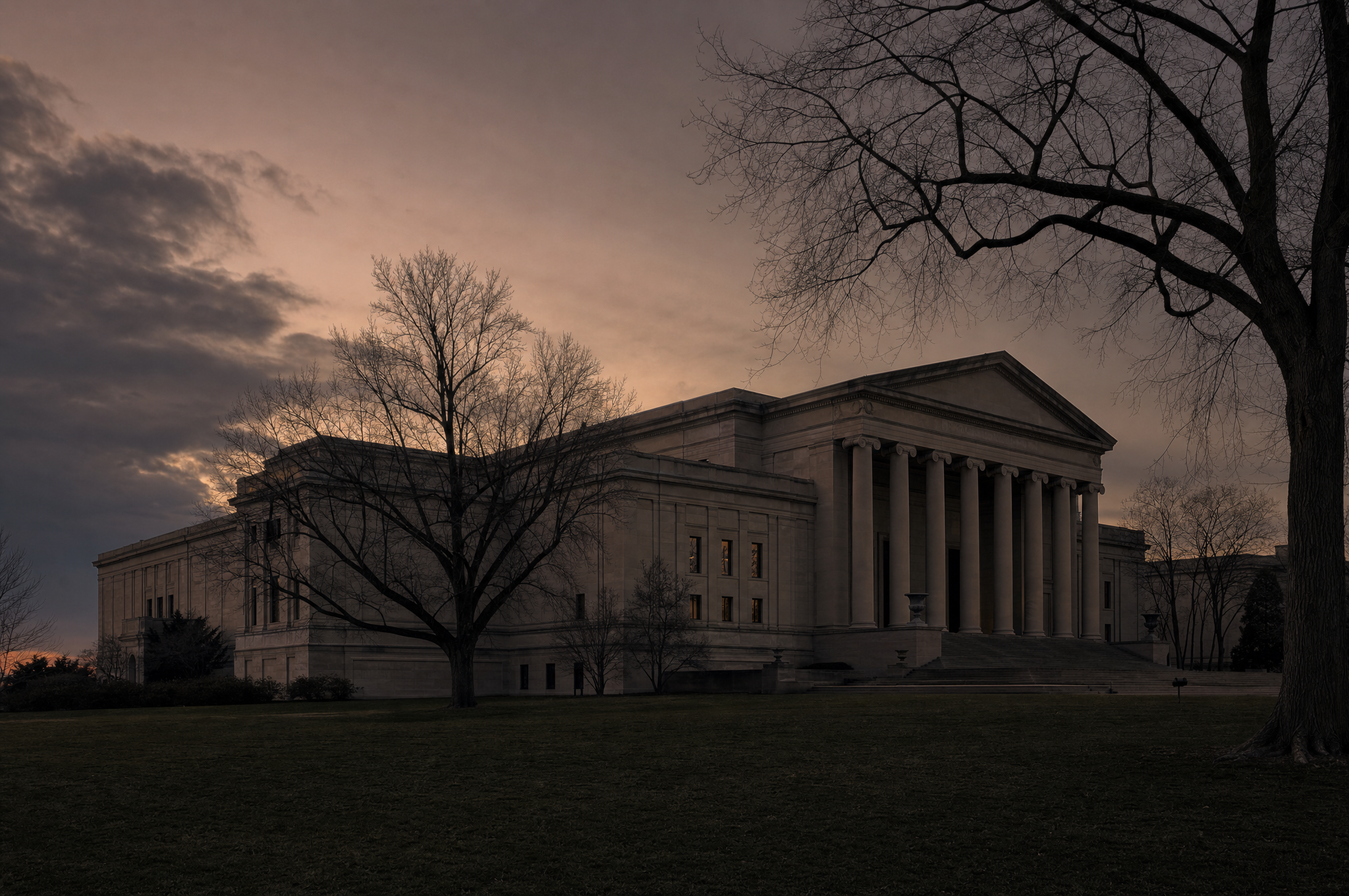 A neoclassical limestone museum building on a wide lawn at dusk, long shallow steps and bare trees