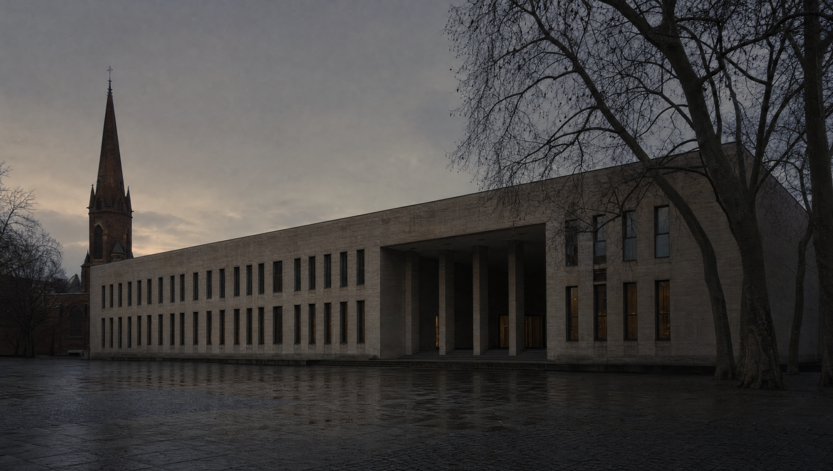 The sober pale stone block of the Kupferstichkabinett at the Kulturforum seen across an empty plaza at dusk, bare winter trees and the brick spire of St Matthew's Church behind