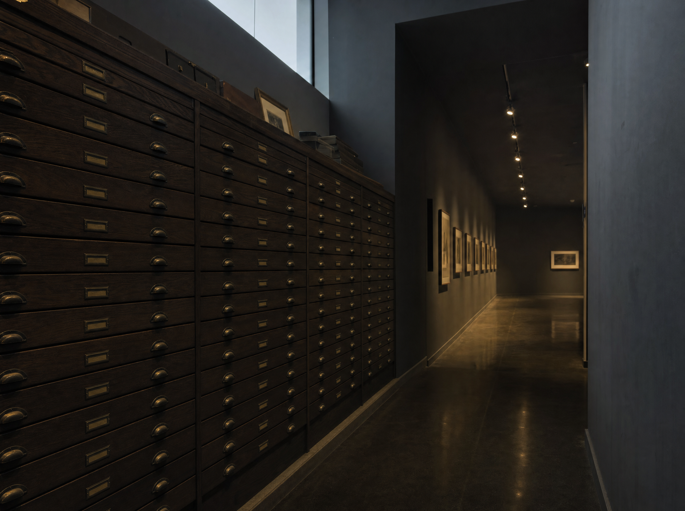 A bank of shallow flat-file drawers along a dark corridor, with a dimly lit exhibition gallery of framed prints beyond