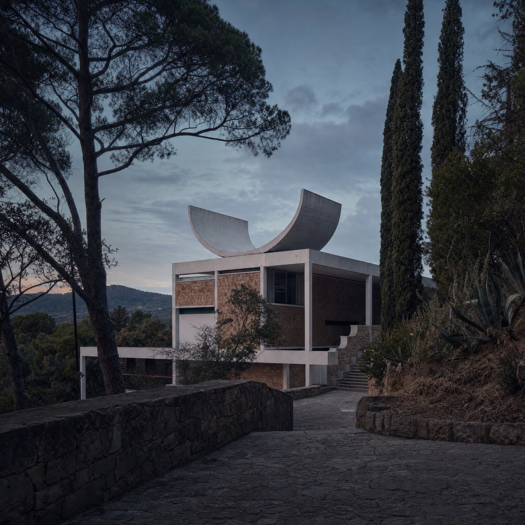 The Fondation Maeght at dusk: rose-ochre stone and white concrete beneath two curved roof sails, pines and cypress on the approach