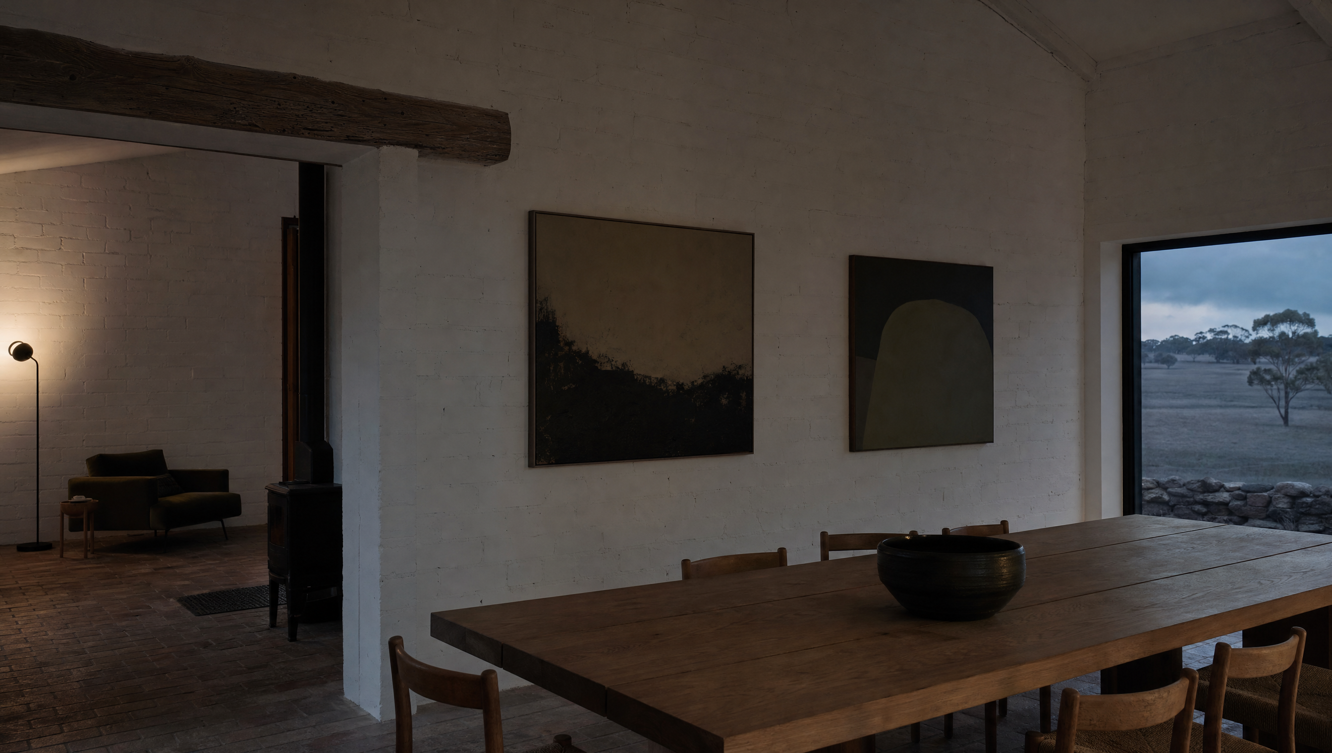 An empty whitewashed farmhouse room with a plain timber table and paintings hung low
