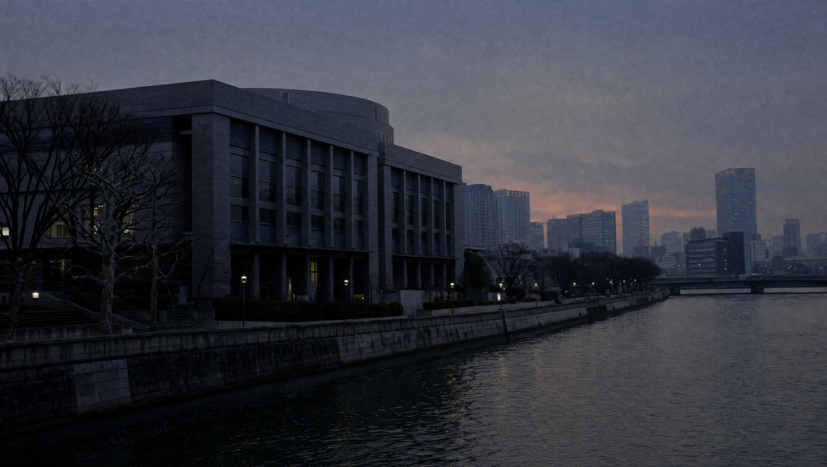 A broad river through a Japanese city at blue hour, stone embankment and a dark granite civic building on the bank
