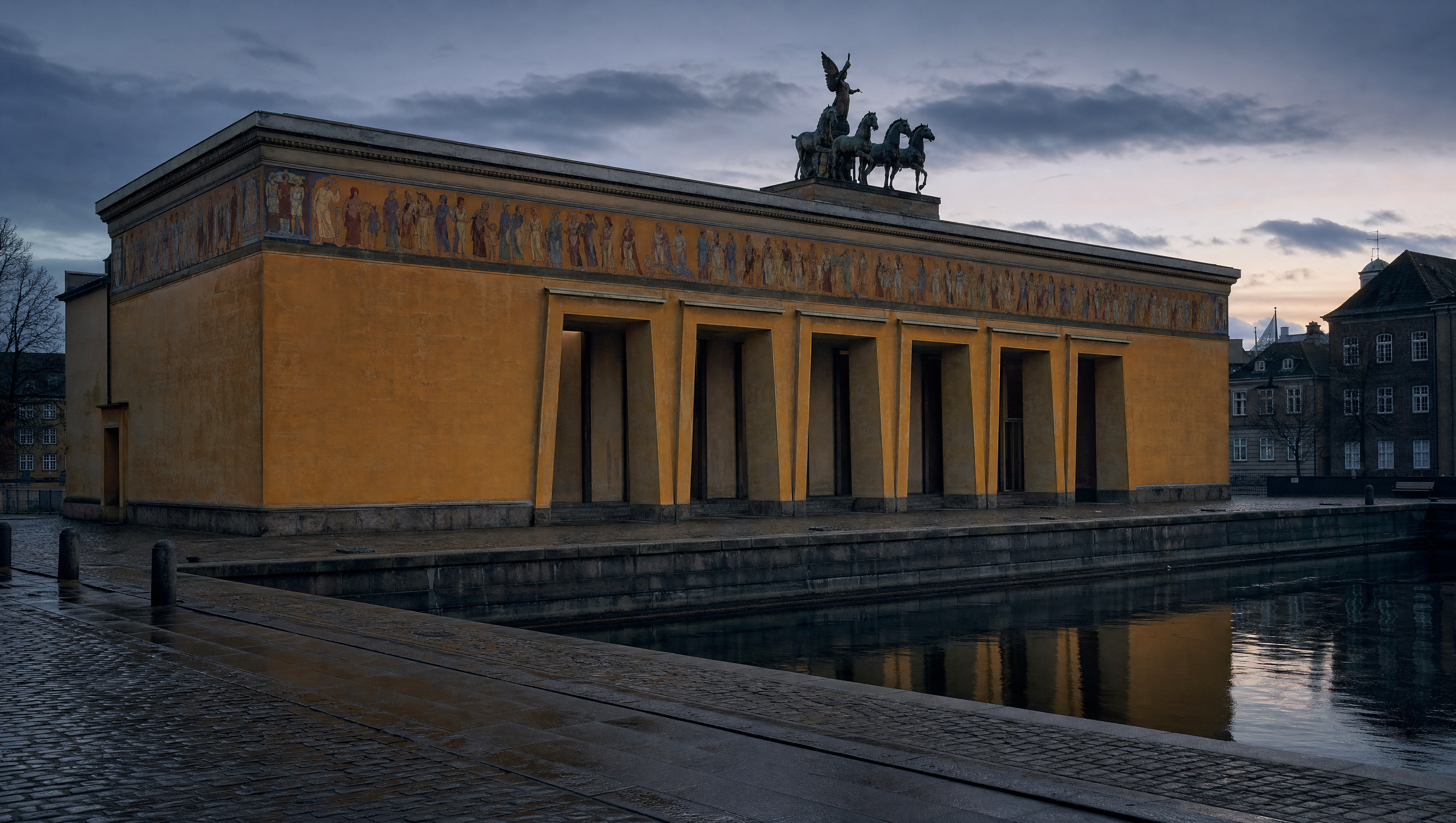 The ochre canal facade of Thorvaldsens Museum at dusk, its tall trapezoidal portals and roofline quadriga