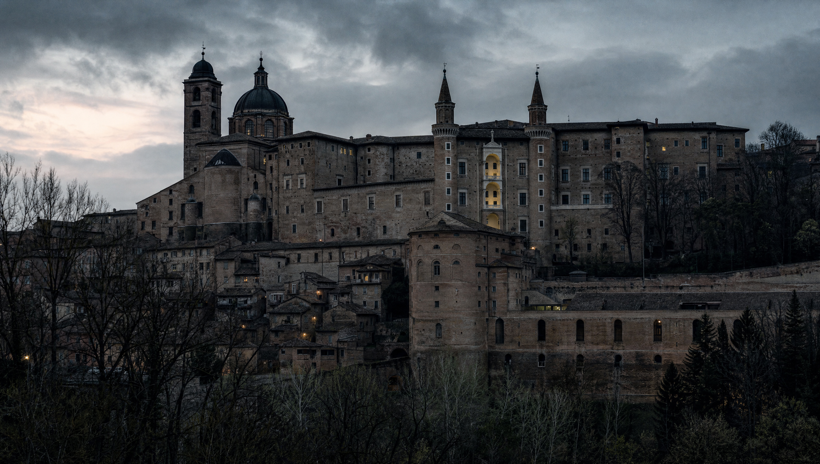 The hill town of Urbino at dusk seen from below, palace towers and brick roofs against a cold sky