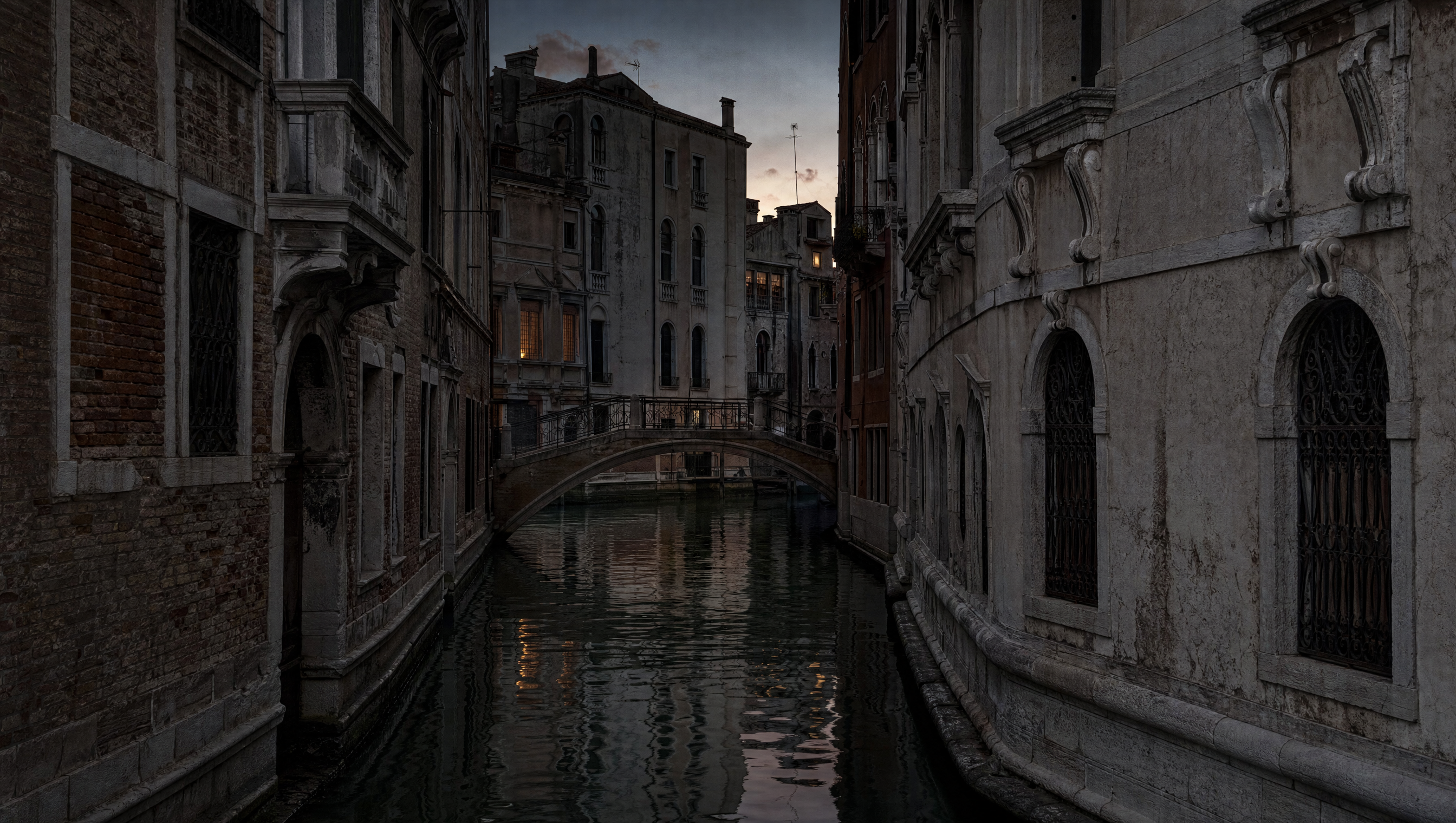 A narrow Venetian canal at dusk in November, brick and stone walls meeting still dark water