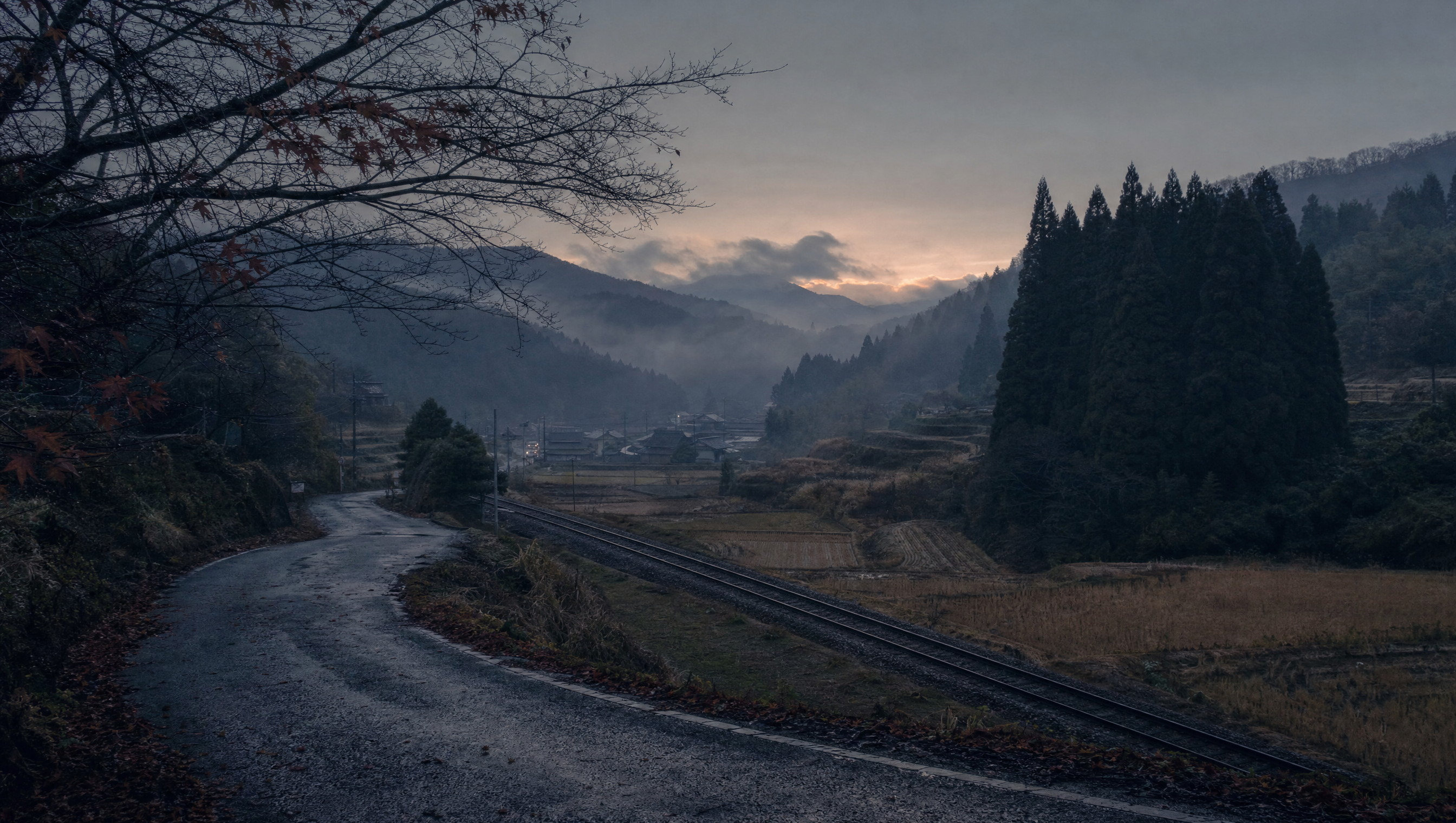 A rural Japanese valley at dusk in late November, terraced fields, pine hills in mist, a single-track railway